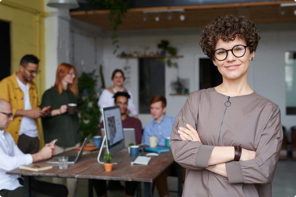 Confident HR professional in a busy modern office with her team behind, representing employee management and self-service software for Australian workplaces