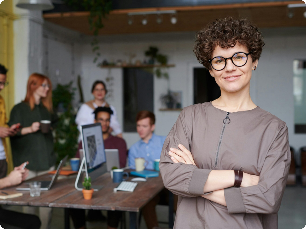 Confident HR manager standing in a busy open-plan office with her team behind, representing employee management software for growing Australian businesses